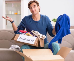 A man seated on the couch lifts hands in apathy as he places clothing in a box labelled "donations"